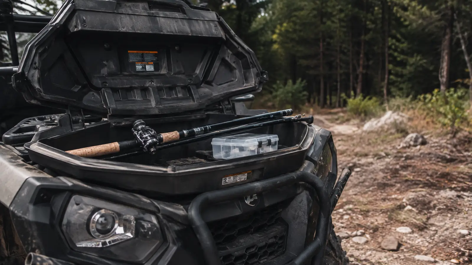 A collapsed telescopic fishing rod resting in the open front storage box of an all-terrain vehicle beside a small tackle box